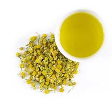 Dried chamomile flowers next to a white cup of yellow chamomile tea.