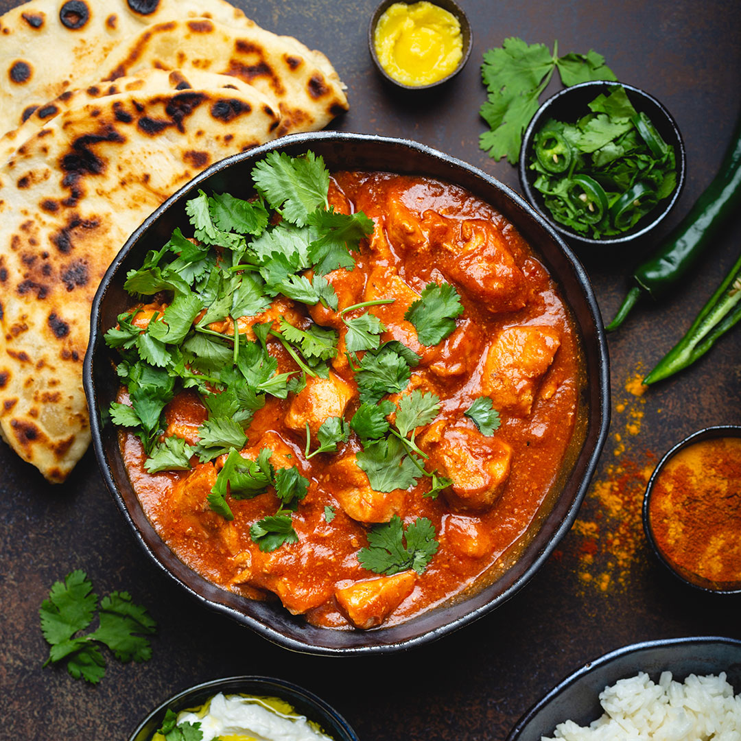 Bowl of tandoori chicken curry with naan bread, chutneys, and garnishes